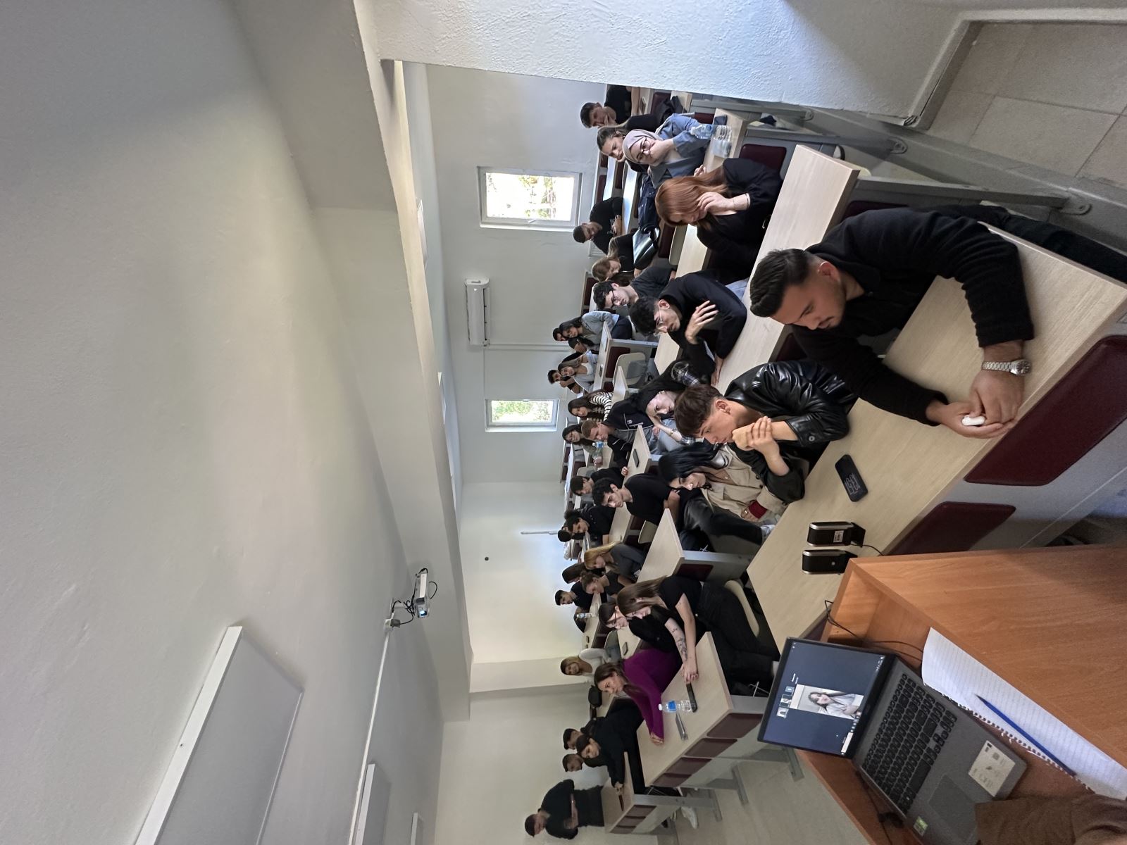 Bu fotoğrafta sınıfın daha geniş bir açıdan görünümü yer alıyor. Sıralarda oturan yaklaşık otuz öğrenci dikkatle öne doğru bakıyor. Çoğu genç, üniversite öğrencisi. Bazıları ellerini çenesine koymuş, bazıları defterlerine yaslanmış şekilde sunumu izliyor. Önde bir masa üzerinde açık bir laptop bulunuyor ve ekranda çevrim içi konuşma yapan bir kadın görülüyor. Sınıf düzenli, öğrenciler yoğun bir dikkatle konuşmacıyı dinliyor.